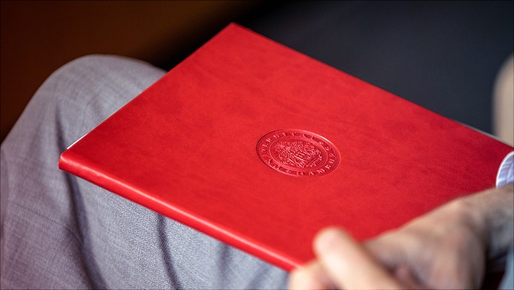 Ein rotes Buch mit Prägung eines Emblems liegt auf dem Bein einer Person.