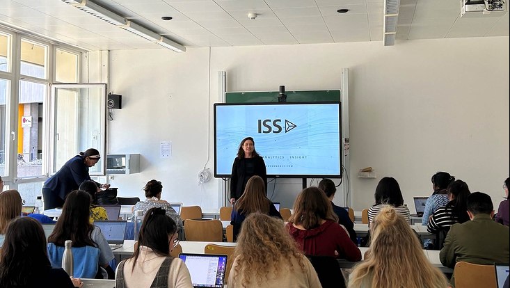 Auf dem Bild sieht man wie Kristina Rüter vor den Studierenden präsentiert und die Präsentation im Hintergrund zu sehen ist.