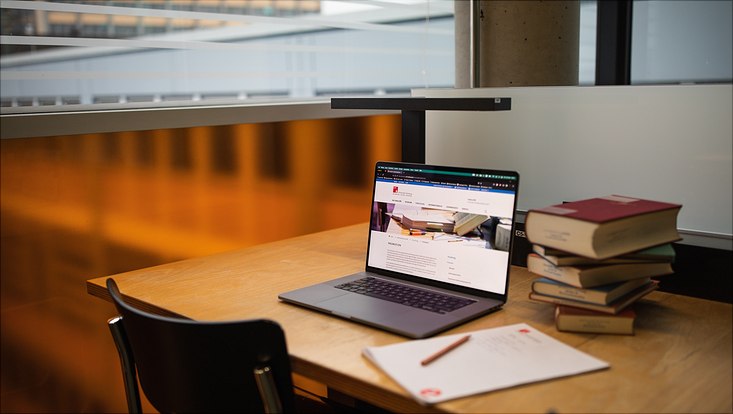 Aufgeklappter Laptop mit einem Bücherstabel rechts daneben und einem UHH-Schreibblock mit Bleistift davor, in der Bibliothek der Rechtswissenschaft