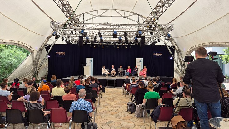 Tagung mit großer Bühne, auf der fünf Podiumsteilnehmer diskutieren, vor einem gefüllten Saal