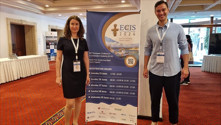 Female (on the right) and male-presenting individual (on the left) stand next to a sign for the 32nd European Conference on Information Systems