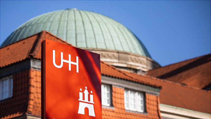 Weißer Schriftzug UHH mit Logo auf roten Viereck vor dem Hauptgebäude der Uni Hamburg, grüne Kuppel vor roten Dächern