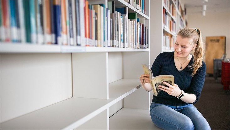 frau sitzt vor buecherregal in der bibliothek und liest buch