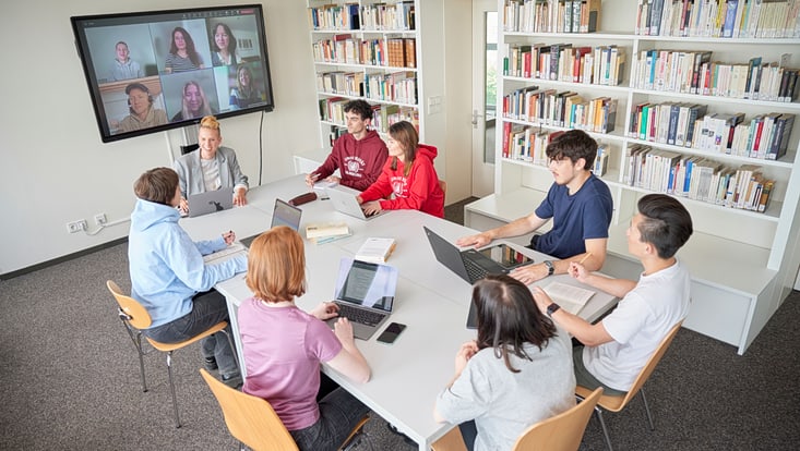 Blick auf eine Gruppe Studierender mit Dozentin um einen Tisch herum, im Hintergrund ein Bildschirm mit Bildern der virtuell Teilnehmenden