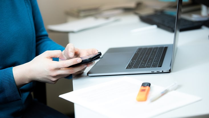 Eine Person sitzt vor einem Laptop und tippt auf ihrem Handy