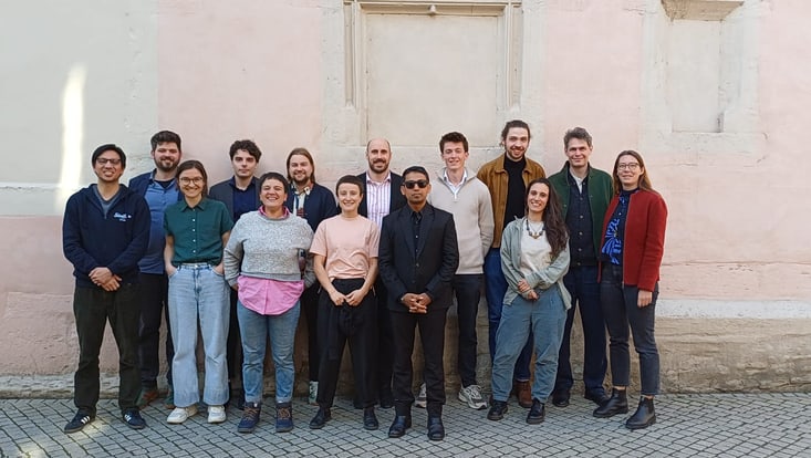 14 Personen stehen als Gruppe in zwei Reihen vor einer Wand. Alle lächeln, sie tragen sportliche Alltagskleidung, dazu Sneaker oder Stiefel.