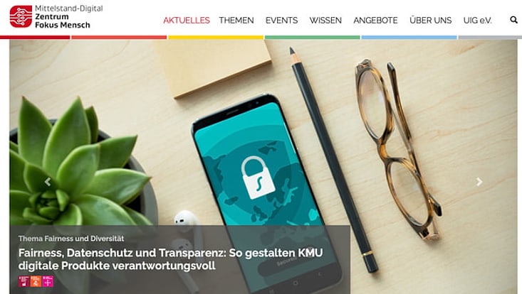 Ausschnitt der Website Mittelstand-Digital Zentrum Fokus Mensch. Abbildung Schreibtisch Brille, Handy, Stift, Kopfhörer, Pflanze. Schrift Thema Fairness und Diversität