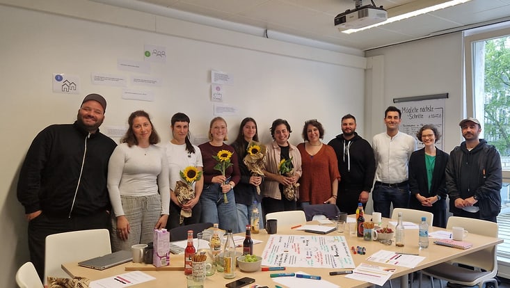Raum der Universität Hamburg. 11 Personen stehen in einer Reihe hinter einem Konferenztisch und schauen in die Kamera.