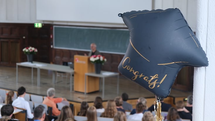im Vordergrund ein Balloon mit "Cangrats!" drauf; Im Hintergrund ist ein Mann auf einer Bühne zu sehen, mehrer Menschen schauen ihm zu
