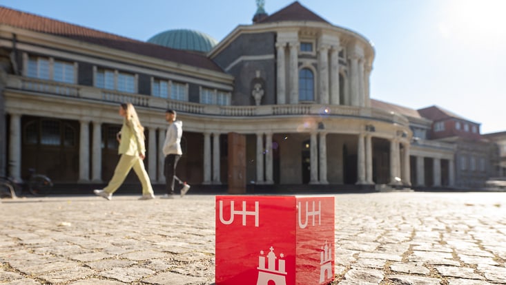 Ein roter UHH-Würfel liegt in der Sonne vor dem Hauptgebäude. Im Hintergrund gehen zwei Menschen.