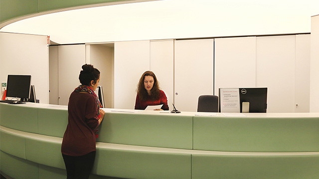 Mitarbeiterin im Studienbüro mit Zettel in der Hand sitzt hinter dem grünen Tresen und Studierende mit hochgesteckter Frisur steht vor dem Tresen