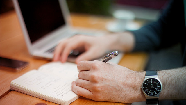 Jemand schreibt etwas in ein Notizheft, ein Laptop liegt daneben