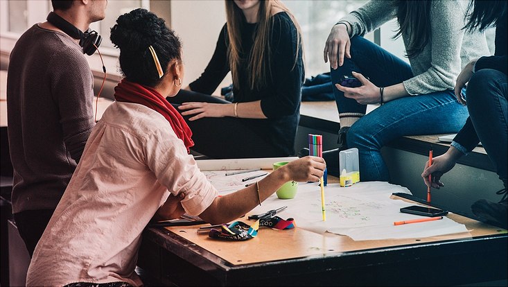 Eine Gruppe von jungen Menschen sitzt um einen Tisch auf dem Arbeitsmaterialien liegen. Ihre Gesichter sind nicht zu erkennen, da sie außerhalb des Bildrands sind.