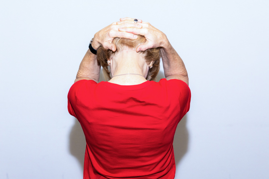 Nacken dehnen Übung zur Augenentspannung: Eigenmassage - Eine Frau ist von hinten zu sehen. Ihre Hände hat sie auf ihren Hinterkopf gelegt.