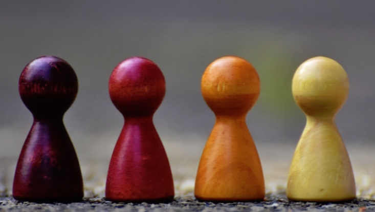 4 Spielfiguren auf einer Linie nebeneinander. Die Figuren haben unterschiedliche,rötliche Farben und stehen auf einem dunklen Untergrund