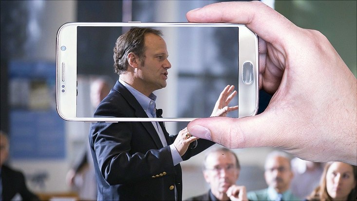 Ein Mann trägt in einem Raum vor Menschen vor und wird dabei mit dem Handy gefilmt.
