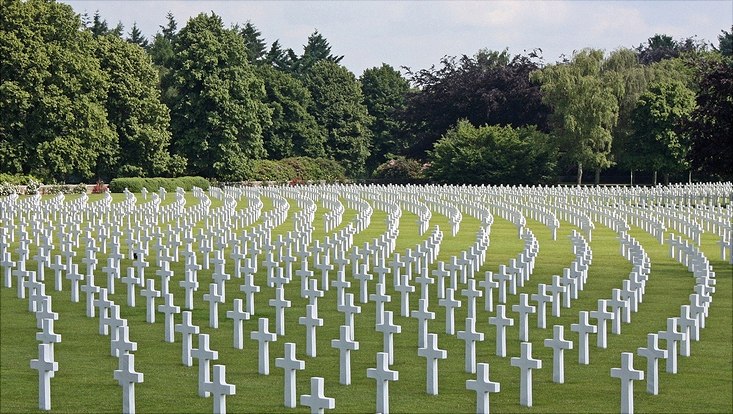 Es ist ein Friedhof im Grünen zu sehen, auf dem unzählige weiße Kreuze aufgestellt sind. Im Hintergrund stehen grüne Bäume.