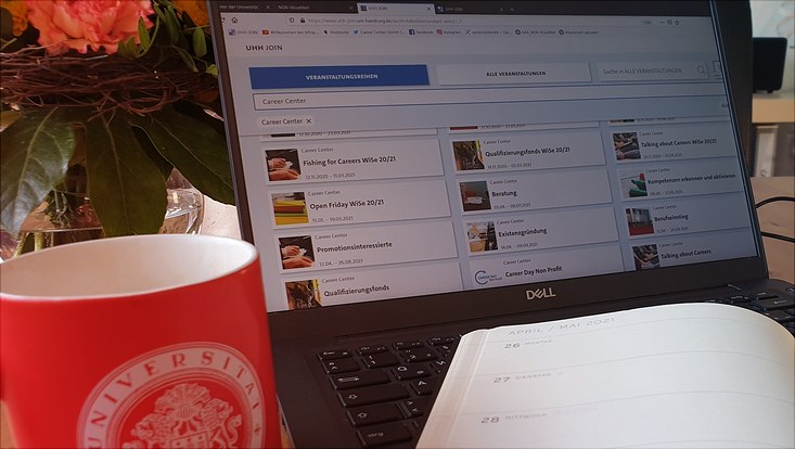 Rote Tasse mit UHH-Logo vor aufgeklapptem Laptop, ein aufgeklappter Kalender auf der Tastatur