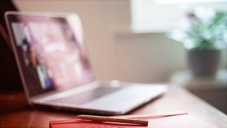 Aufgeklappter Laptop, der BIldschirm ist unscharf aber man sieht ein laufendes digitales Meeting. Vor dem Laptop ist ein orangenes Notizheft mit Stift auf dem Tisch.