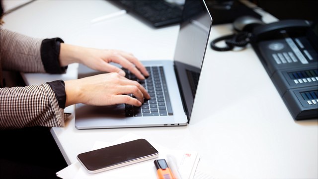 Frau mit hochgekrempelten Ärmeln arbeitet am Laptop, neben ihr liegt ein Smartphone