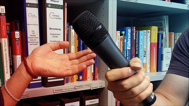 Eine Hand hält ein Mikrofon, eine zweite Hand einer Interviewpartnerin wird in einer Geste mit nach oben geöffneter Handfläche daneben gehalten.