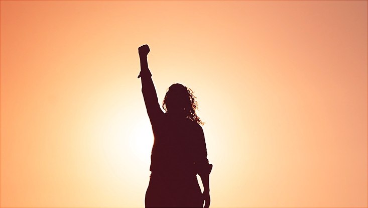 Eine Frau steht mit erhobener Faust vor der Sonne. Es ist nur ihre Silhouette zu sehen.