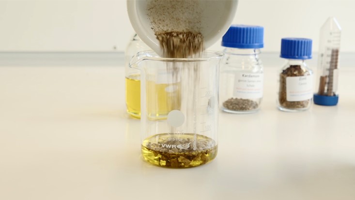 Startscreen für den Film „Kinder-Uni 2021: Parfümherstellung in der Antike“ – ein gemörstertes Pulver wird in ein mit Öl gefülltes Glasgefäß gefüllt.