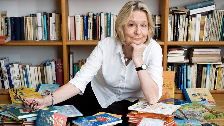 Frag Kirsten Boie! Kirsten Boie sitzt vor einem Bücherregal. Vor ihr liegen zahlreiche Bücher durcheinander