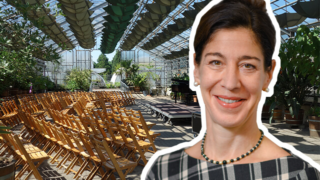 Im Vordergrund ist Prof. Dr. Anne van Aaken abgebildet und im Hintergrund ein Gewächshaus des Botanischen Gartens in Hamburg. Darin stehen Holzstühle und eine kleine Bühne.