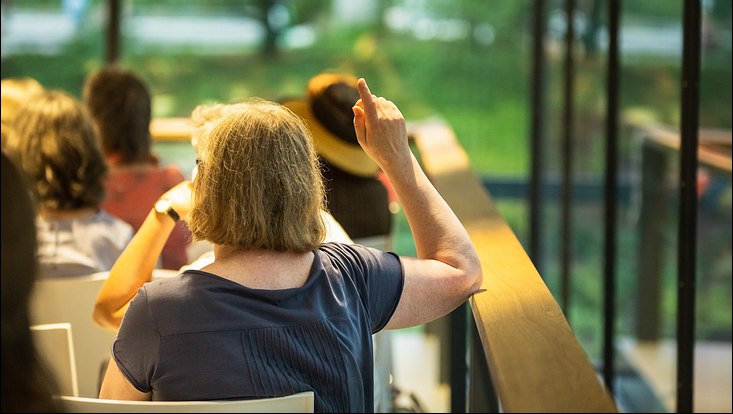 Viele Menschen in einem Raum in einer Workshops-Situation. Eine Frau hebt die Hand, um zu sprechen