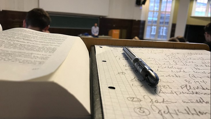 Ein Stift liegt auf mitgeschriebenen Notizen in einem Hörsaal.
