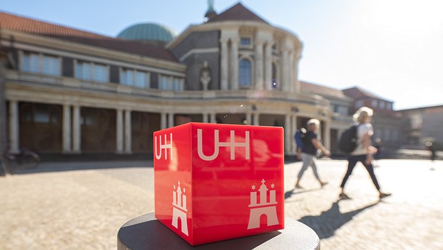 Ein roter Würfel mit der Aufschrift "UHH" steht vor dem Hauptgebäude der Universität Hamburg. Zwei Personen laufen durch das Bild.
