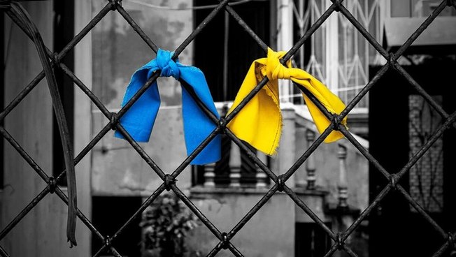 Titelbild der Ausstellung: ein Stück gelber und ein Stück blauer Stoff sind an einen Zaun geknotet