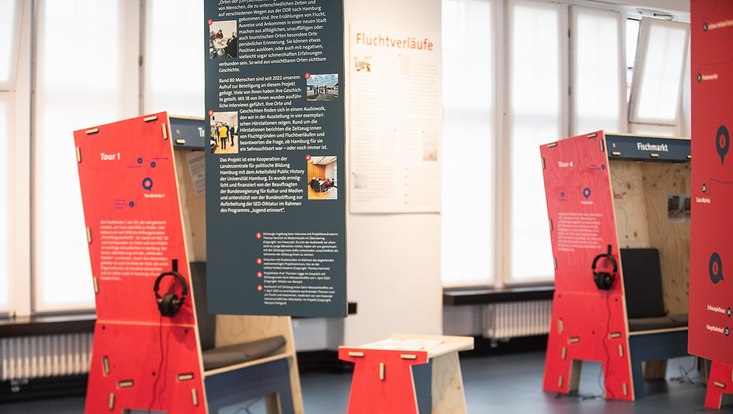 Foto von der Ausstellung "Orte der Unsichtbarkeit": Blick auf eine Informationstafel sowie auf zwei Audiostationen, an denen Kopfhörer hängen.