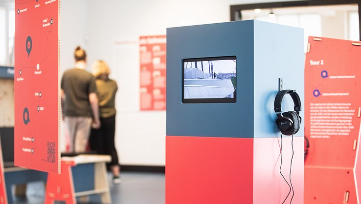 Foto von der Ausstellung "Orte der Unsichtbarkeit": Blick auf eine Video-Steele. Auf einem kleinen Monitor ist der Ausschnitt einer Brücke zu sehen. An der Steele hängt ein Kopfhörer. Im Hintergrund sind zwei Besucher:innen der Ausstellung zu sehen.