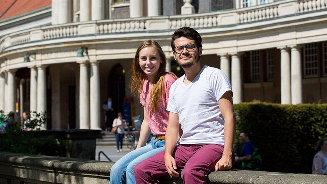 Two students sit in the sun in front of Universität Hamburg’s Main Building.
