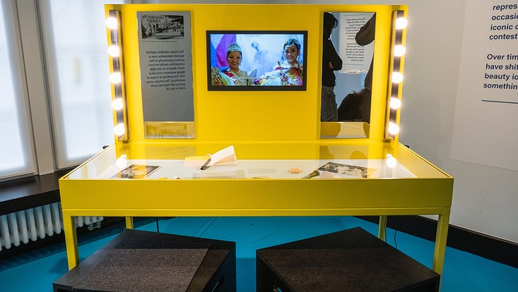Sonderausstellung „Traces of Empires in the Philippines“ Foto eines beleuchteten gelben Spiegeltisches. Zwischen den beiden Spiegeln auf der Rückwand ist ein Monitor eingebaut, der einen Film abspielt.