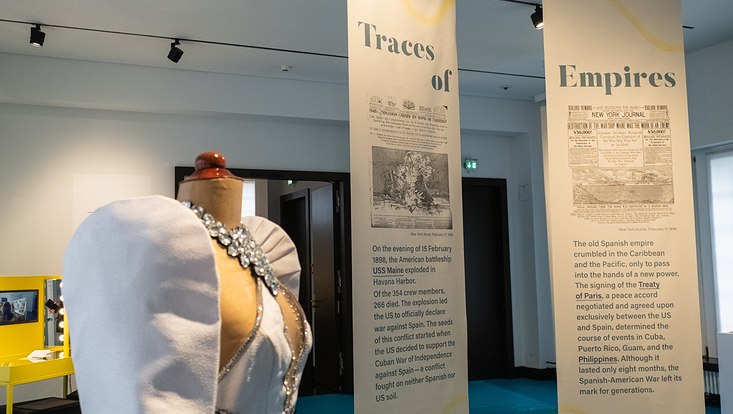 Sonderausstellung „Traces of Empires in the Philippines“ Rechts hängen zwei längliche Poster mit jeweils einem Bild und einem Text von der Decke herunter. Links steht eine Kleiderpuppe ohne Kopf in einem weißen Kleid und einer Edelsteinkette. Links hinter der Puppe ist ein gelber Spiegeltisch aufgebaut.