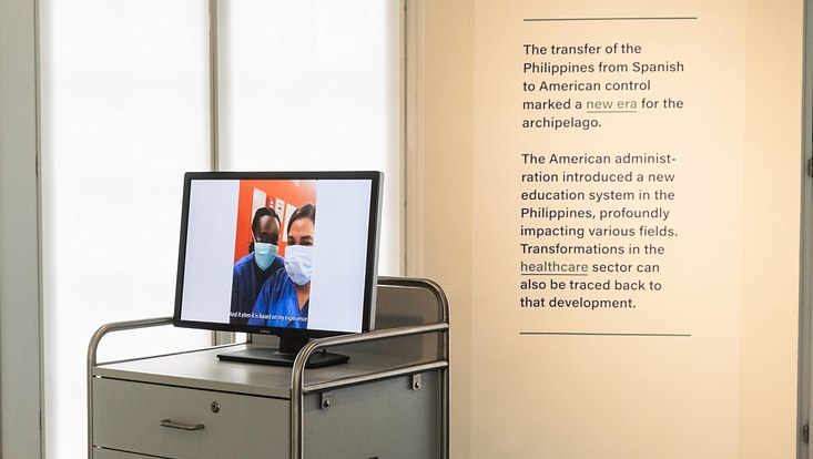 Sonderausstellung „Traces of Empires in the Philippines“ Rechts ist ein Text auf einer Leinwand abgebildet und links daneben steht ein Schrankwagen aus dem Krankenhaus, auf dem ein Bildschirm platziert ist. Auf dem Bildschirm wird ein Bild von zwei Pflegekräften gezeigt.