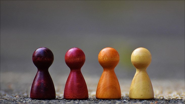 4 Spielfiguren auf einer Linie nebeneinander. Die Figuren haben unterschiedliche,rötliche Farben und stehen auf einem dunklen Untergrund