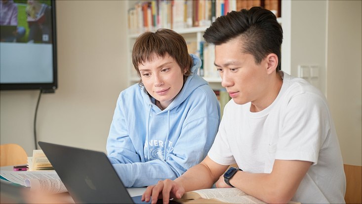 Zwei Studierende sitzen an einem Tisch und schauen gemeinsam auf einen Laptop.