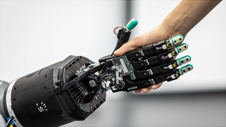 Eine menschliche Hand drückt eine Roboterhand. Die Roboterhand hat bewegliche Finger und freiliegende Drähte.