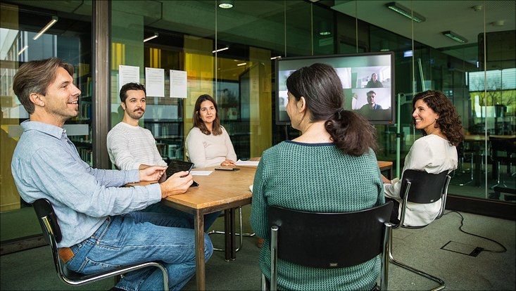 Fünf Personen sitzen um einen Tisch in einem Besprechungsraum, zwei Personen führen eine Videokonferenz, die auf einem Bildschirm angezeigt wird. Der Raum hat Glaswände und im Hintergrund hängen einige Papiere.