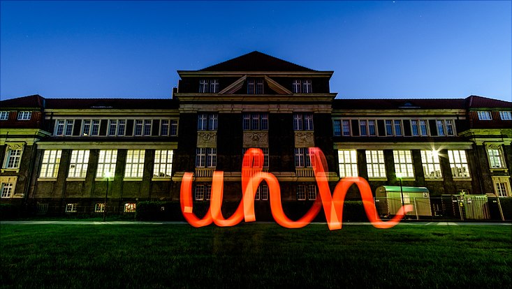 Das Hauptgebäude der Uni Hamburg bei Nacht. Davor ein neonroter UHH-Schriftzug.