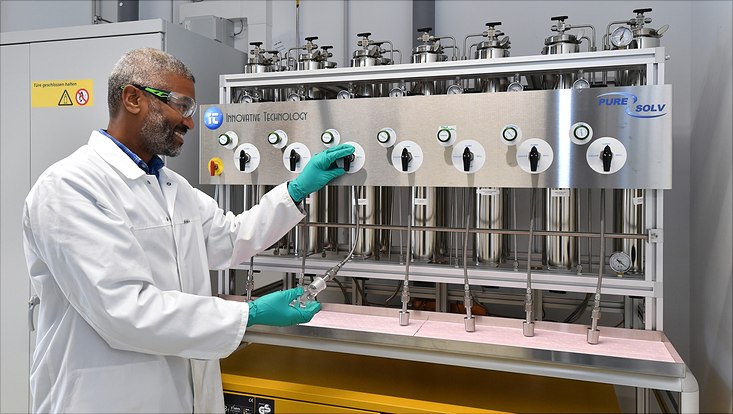 Eine Person in Laborkittel und Handschuhen bedient eine Maschine mit mehreren Messgeräten und Ventilen in einer Laborumgebung.