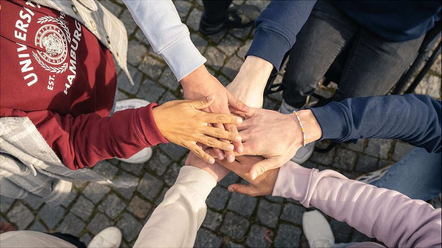 Sechs Personen stehen im Kreis, strecken die Arme aus und legen ihre Hände aufeinander. Die Hände haben unterschiedliche Hauttöne.