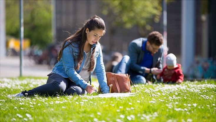 Eine Studentin sitzt auf einer Wise und lernt. im Hintergrund sitzt ein Mann mit einem Kleinkind.