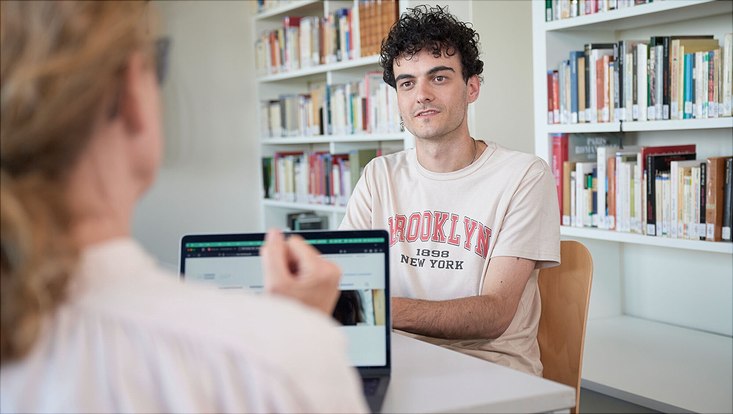 Ein Mann sitzt in einer Beratungssituation an einem Tisch gegenüber einer anderen Person, die nur im Anschnitt von hinten zu sehen ist.