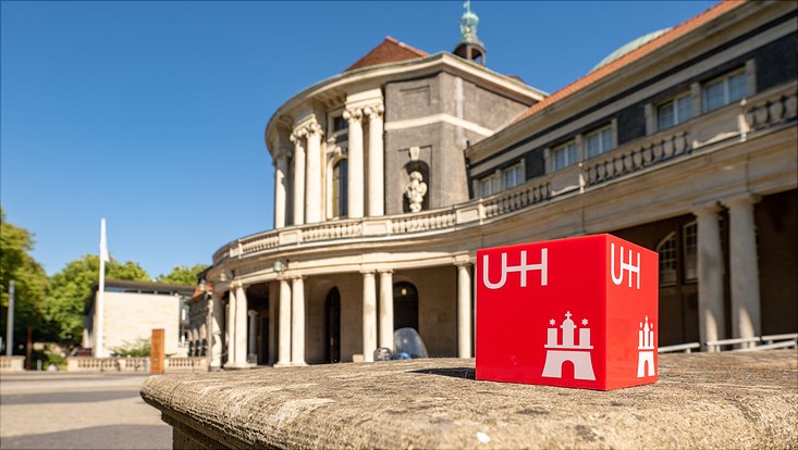 Roter Würfel mit der Aufschrift UHH und im Hintergrund das Hauptgebäude der Universität