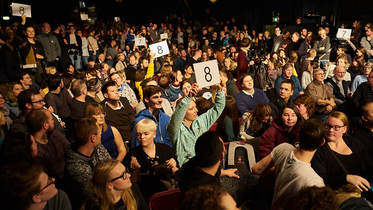 Publikum bei einem Scienceslam, manche halten Schilder mit Punktebewertungen hoch